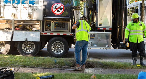 Workers in high-visibility vests using an industrial vacuum truck for ground excavation.