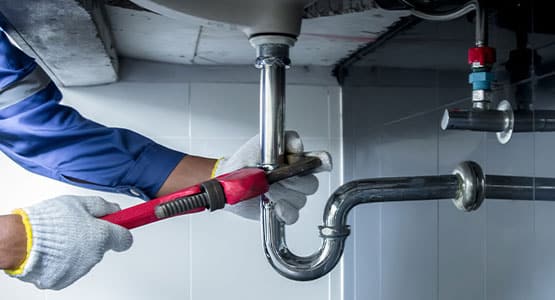 Plumber using wrench to fix under-sink pipe, wearing gloves and blue shirt.