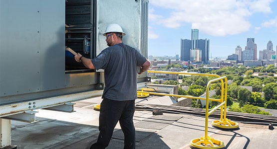 A worker in a hard hat inspects rooftop HVAC equipment with a city skyline in the background.