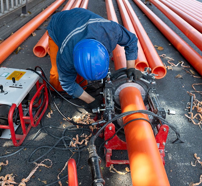 A worker in blue helmet assembles orange pipes with a fusion machine outdoors.