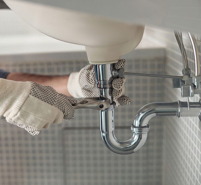 A plumber fixing a sink pipe with a wrench in a tiled bathroom.