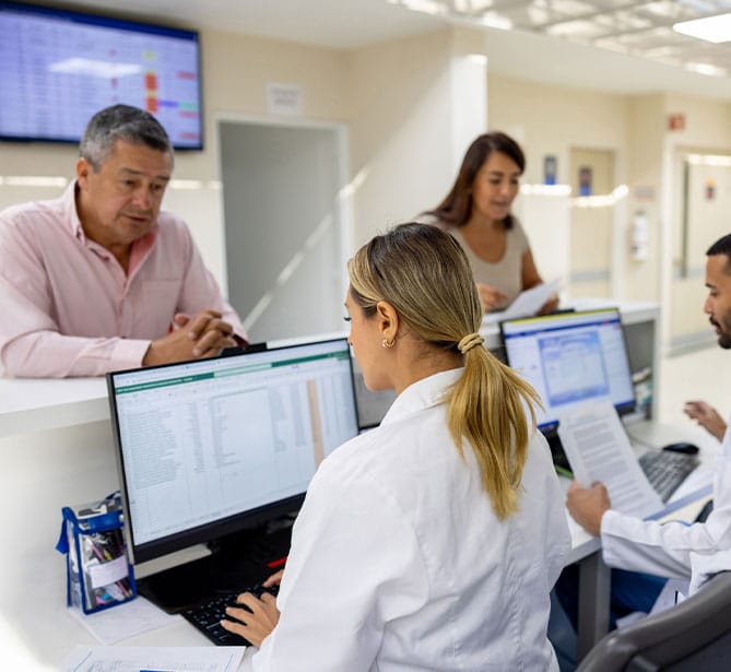 People at hospital reception desk interacting with staff, computers on desks.