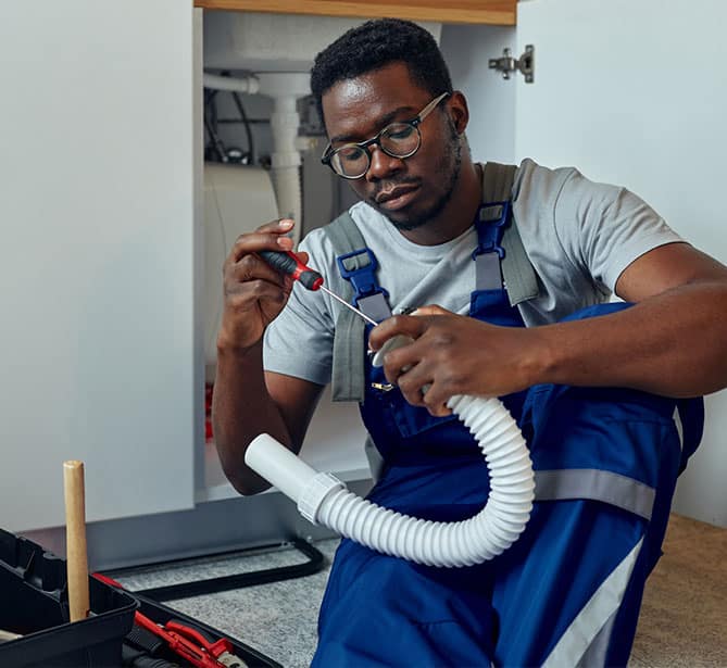 A plumber fixing a pipe under a sink, using a screwdriver.