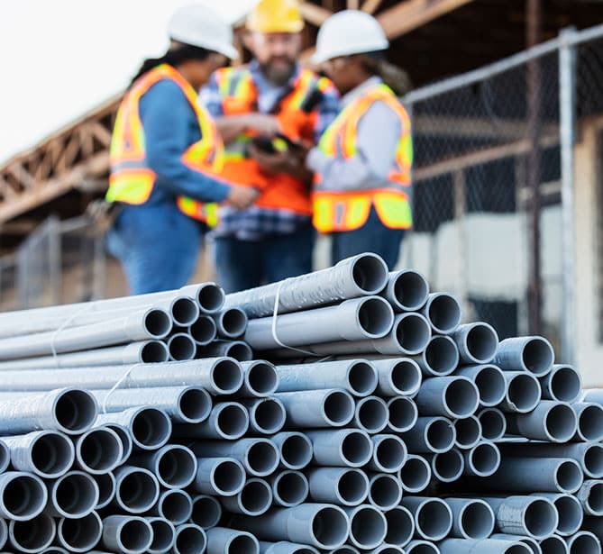 Pipes stacked in the foreground with construction workers in safety gear discussing in the background.