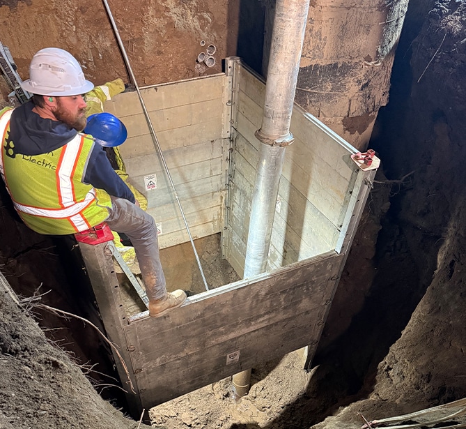 Worker in safety gear inspecting a utility trench with a metal support structure.