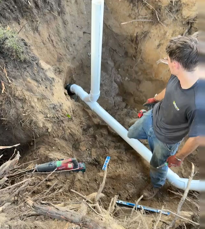 A man works on installing a PVC pipe in a trench with tools nearby.