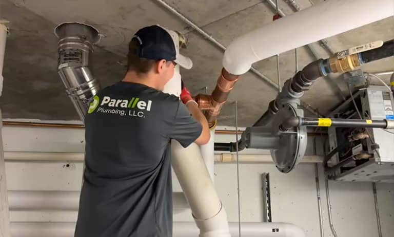 A plumber from Parallel Plumbing works on pipes in a building's utility room.