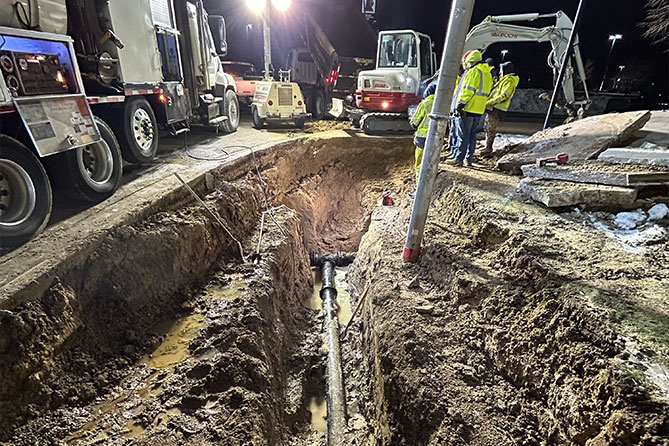 Excavation site with construction equipment and workers in safety vests installing underground pipeline at night.