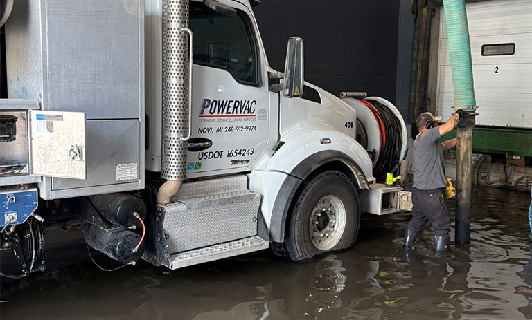 A man operates vacuum truck equipment in a flooded area near a building.