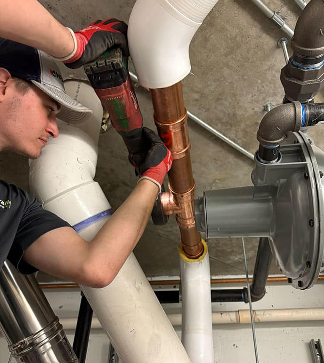 A plumber working on pipes with a power tool in a basement setting.