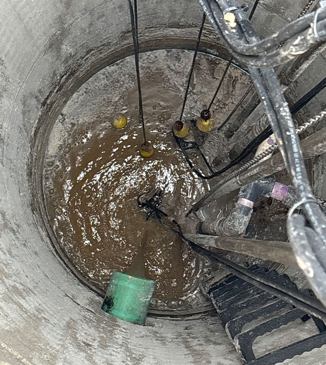 Concrete pit with pipes, cables, and murky water, viewed from above.