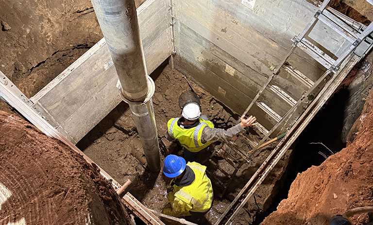 Two construction workers in safety gear working in a deep trench with ladders and soil.