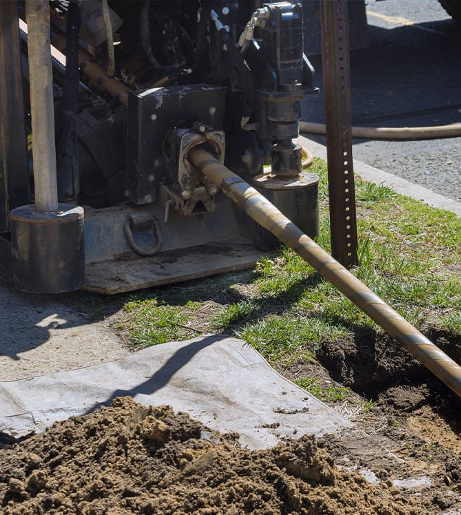 Pipe drilling machine in action on grassy area near sidewalk, surrounded by dirt and equipment.